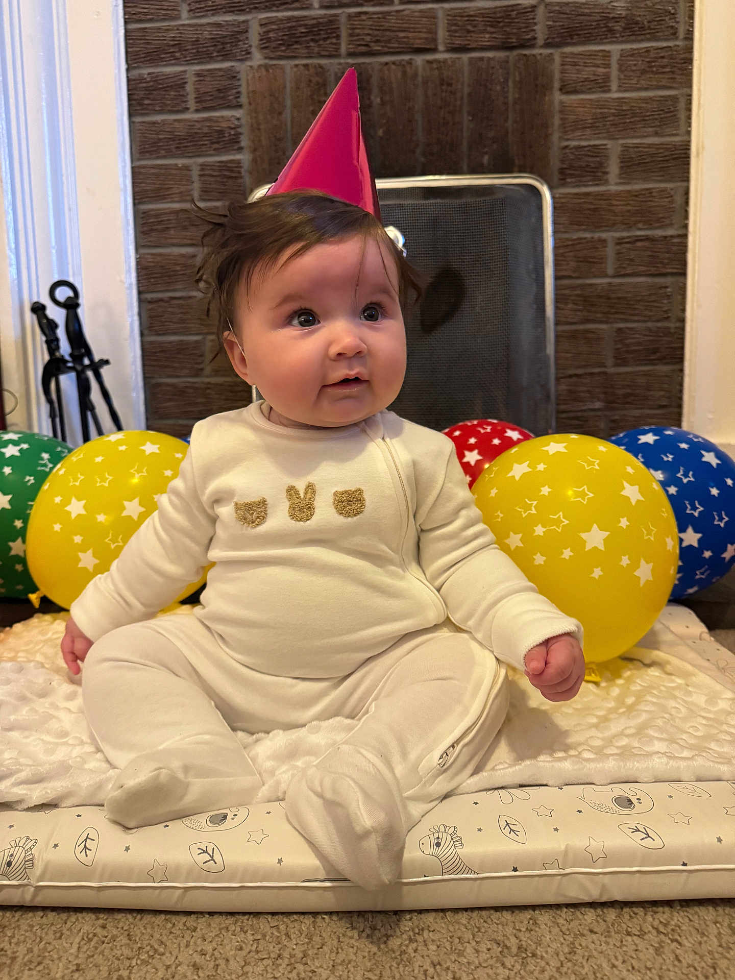 Frankie is registered to the contest to win money with this photo: baby, party_hat, balloons, indoor, carpet, blanket, cushion, cute, child, white_clothing, celebration, birthday, infant, plush, face, sitting, floor, headwear, smiling, decor