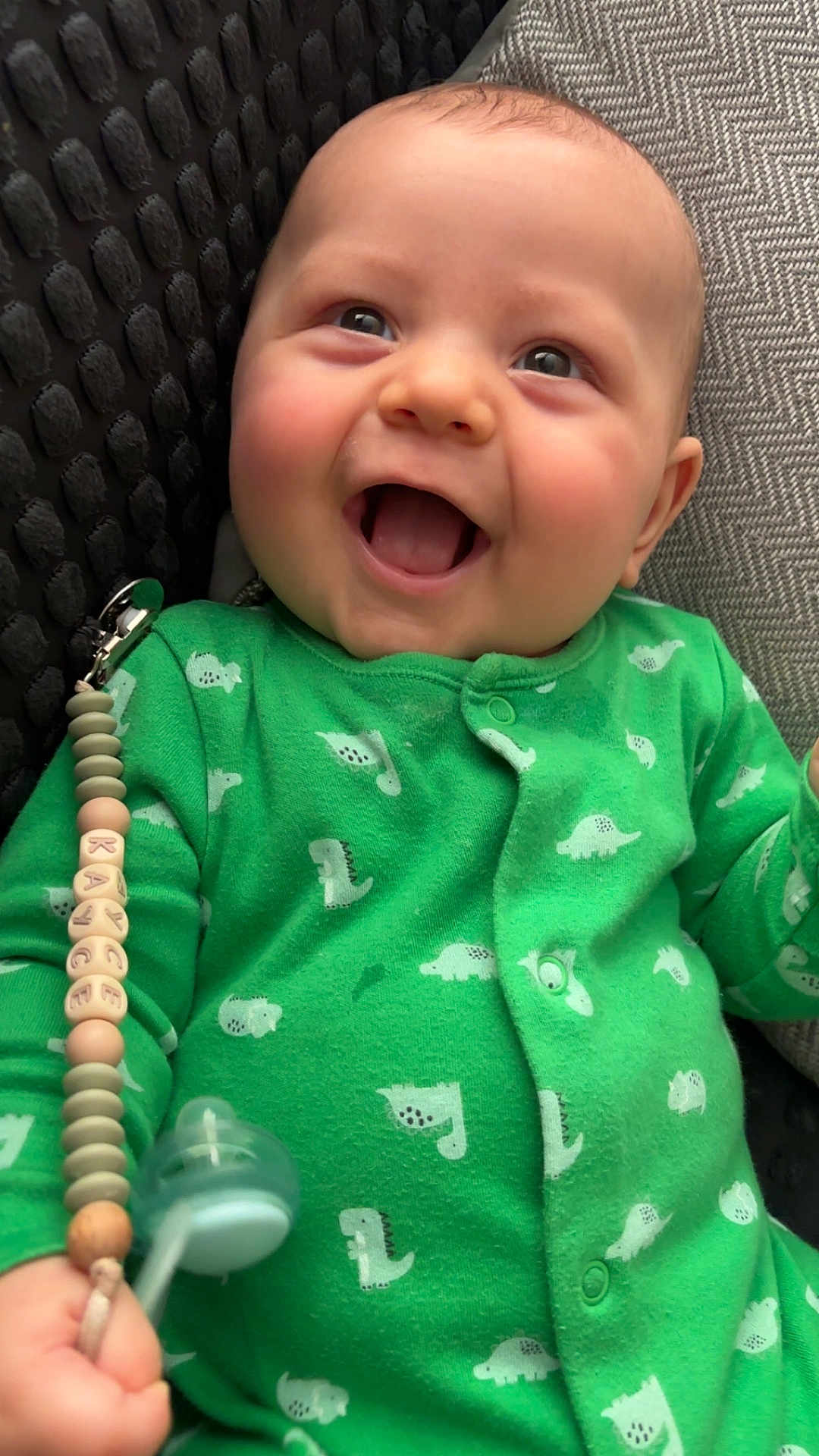 Kayce joined the competition — help win amazing prizes! baby, child, green_onesie, dinosaur_print, pacifier, smiling, happy, infant, cute, clothing, face, hand, indoor, portrait, young_child, comfort, texture, closeup, joy, baby_accessory