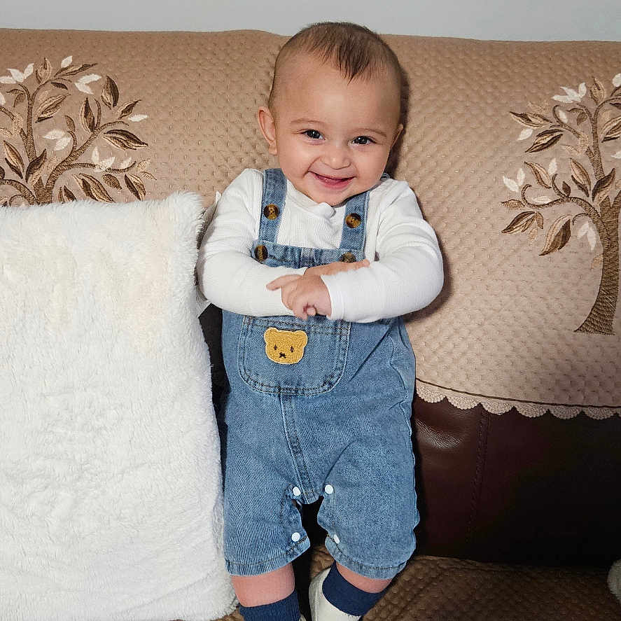Kahan is registered to the contest to win money with this photo: baby, child, couch, cozy, cushion, cute, denim_overalls, fabric, face, footwear, happy, home, indoor, infant, person, portrait, smiling, socks, standing, white_pillow