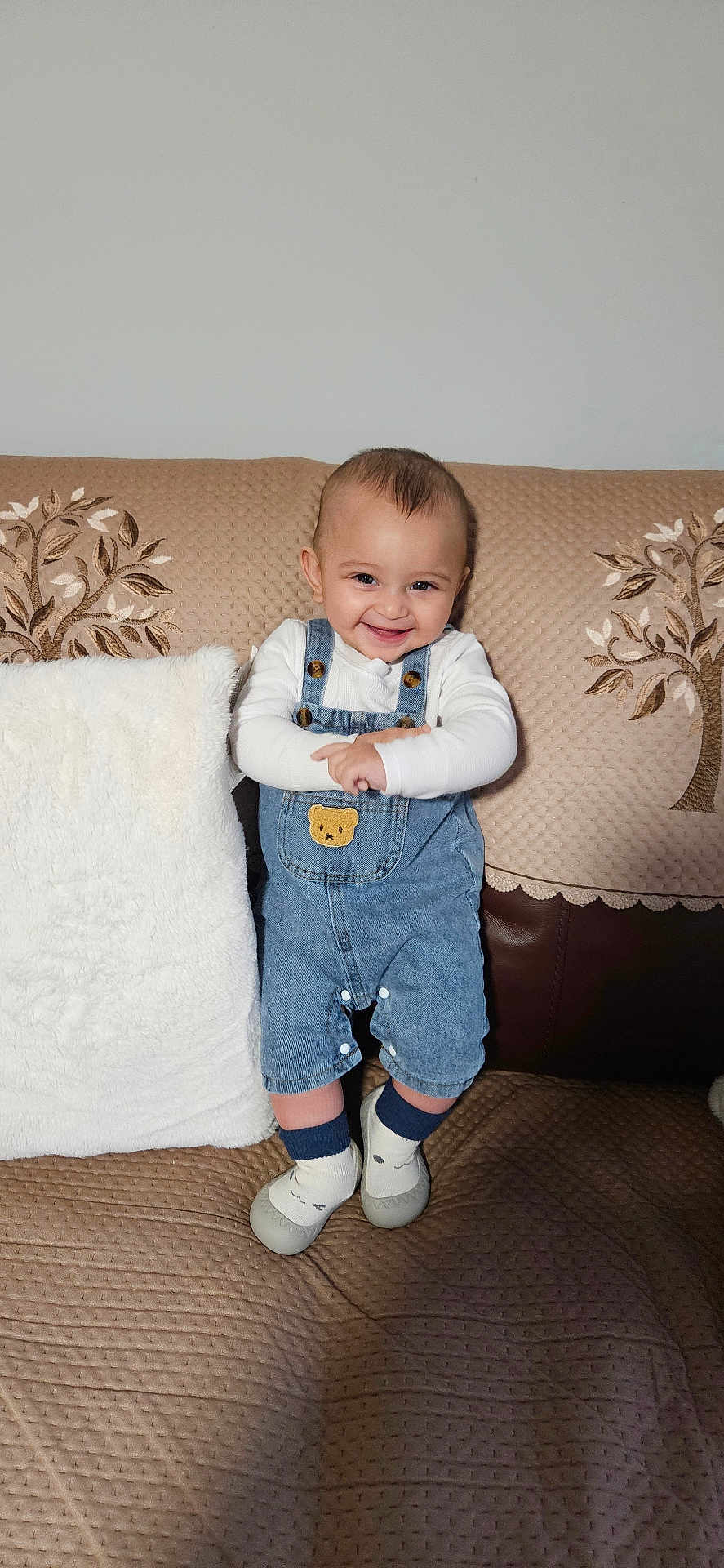 Kahan is registered to the contest to win money with this photo: baby, child, smiling, denim_overalls, couch, cushion, white_pillow, indoor, happy, cute, face, person, footwear, socks, home, portrait, infant, standing, cozy, fabric