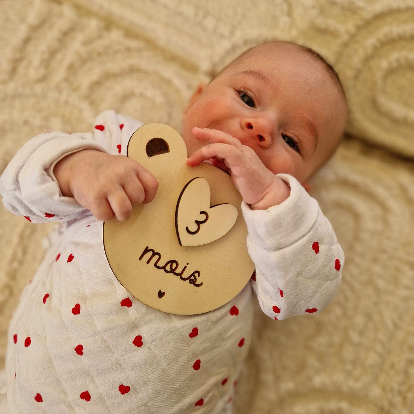 Mia participe au concours pour gagner de l'argent avec cette photo : baby, blanket, child, close_up, cute, face, hand, happy, indoors, infant, lying_down, person, plaque, portrait, red_hearts, smiling, soft_texture, white_clothing, wooden_toy, young