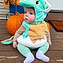 baby, child, costume, dinosaur_costume, pumpkin, autumn, fall, leaves, porch, wooden_floor, cute, plush, sitting, outdoor, seasonal, holiday, face, clothing, sock, expression