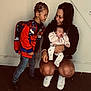 child, baby, woman, indoor, backpack, braids, sneakers, jeans, sweatshirt, holding, sleeping, smiling, curious, family, floor, wall, crouching, affection, casual, portrait