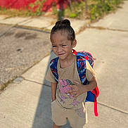 Alexander joined the competition — help win amazing prizes! child, boy, backpack, shorts, tshirt, sneakers, sidewalk, outdoor, sunlight, hair_tied, dinosaur_graphic, casual_clothing, young_child, urban, daytime, concrete, blurred_background, red_wall, greenery, person