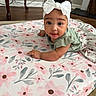 baby, infant, child, headband, bow, blanket, floral_pattern, floor, wooden_table, curly_hair, green_top, skirt, indoors, carpet, face, looking_at_camera, cute, toddler, person, portrait