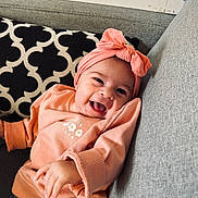 Rose a rejoint le concours — aidez-le/la à gagner de superbes lots ! baby, smiling, headband, bow, peach_sweater, couch, gray_couch, happy, infant, child, portrait, indoors, person, cute, casual_clothing, relaxed, cozy, cheerful, seated, young_child