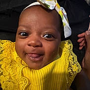 Zuri joined the competition — help win amazing prizes! baby, child, smile, yellow_dress, headband, tongue_out, close_up, portrait, happy, infant, face, hand, clothing, lace, bright_colors, cute, young_child, indoor, person, dark_background