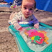 Eddie is registered to the contest to win money with this photo: baby, blanket, blue_background, child, closeup, colorful, curious, cute, face, hands, indoor, infant, lying_down, pajamas, person, play, sea_creatures, soft_texture, toy, water_mat