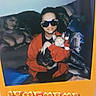 person, cat, sunglasses, red_jacket, couch, blanket, pillow, muscular_man_pillow, calico_cat, indoor, smile, photograph, magnet, pizza_pattern, text_overlay, wall, home, cozy, seated, holding_cat