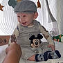 baby, child, smiling, cap, overalls, mickey_mouse, plush_toy, hand, mobile, stars, clouds, curtain, pattern, white_surface, socks, happy, indoor, person, cute, supporting_hand