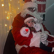 Célina -Marie a rejoint le concours — aidez-le/la à gagner de superbes lots ! baby, child, santa_claus, christmas, holiday, festive, lights, costume, beard, glasses, red_clothing, white_fur, indoor, decorations, person, seated, thumb_sucking, heart_emoji, wall_art, chair
