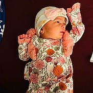 Mirha is registered to the contest to win money with this photo: baby, infant, child, floral_clothing, hat, pink, orange, ruffles, lying_down, maroon_background, newborn, cute, portrait, small_hands, face, clothing, apparel, headwear, looking_sideways, soft_lighting