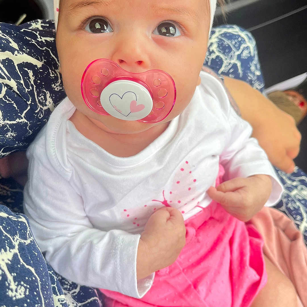Inaya a rejoint le concours — aidez-le/la à gagner de superbes lots ! baby, car_interior, child, closeup, cute, expression, eyes, hand, headband, holding, infant, pacifier, patterned_fabric, person, pink_skirt, portrait, seated, skin, white_clothing, young