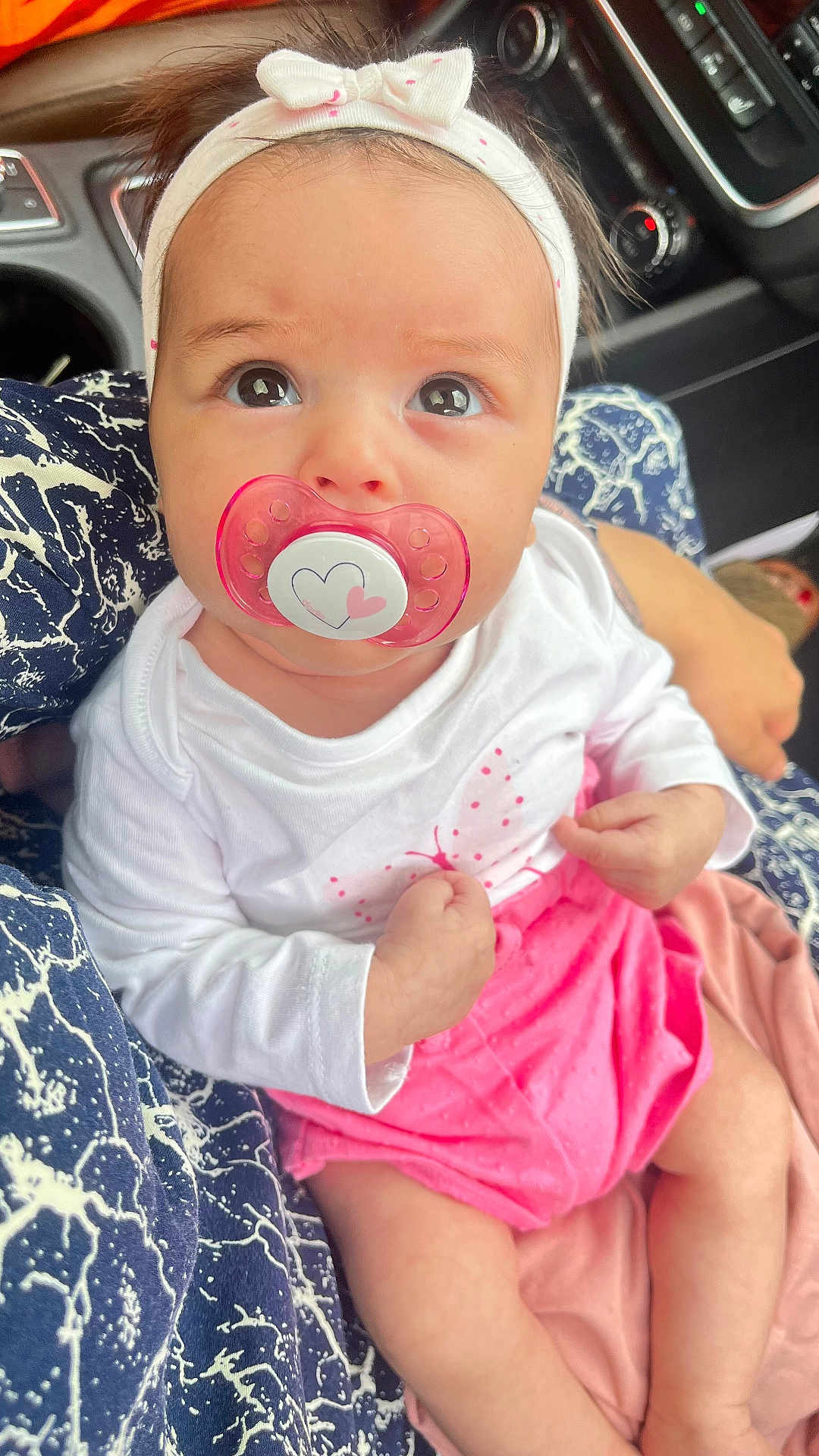 Inaya a rejoint le concours — aidez-le/la à gagner de superbes lots ! baby, pacifier, headband, white_clothing, pink_skirt, infant, person, closeup, car_interior, seated, hand, patterned_fabric, cute, child, portrait, young, skin, eyes, expression, holding