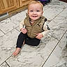 toddler, child, smiling, sitting, indoor, kitchen, floor, tile, marble, wooden_cabinets, pet_crate, clothing, vest, shirt, pants, barefoot, portrait, happy, face, photography