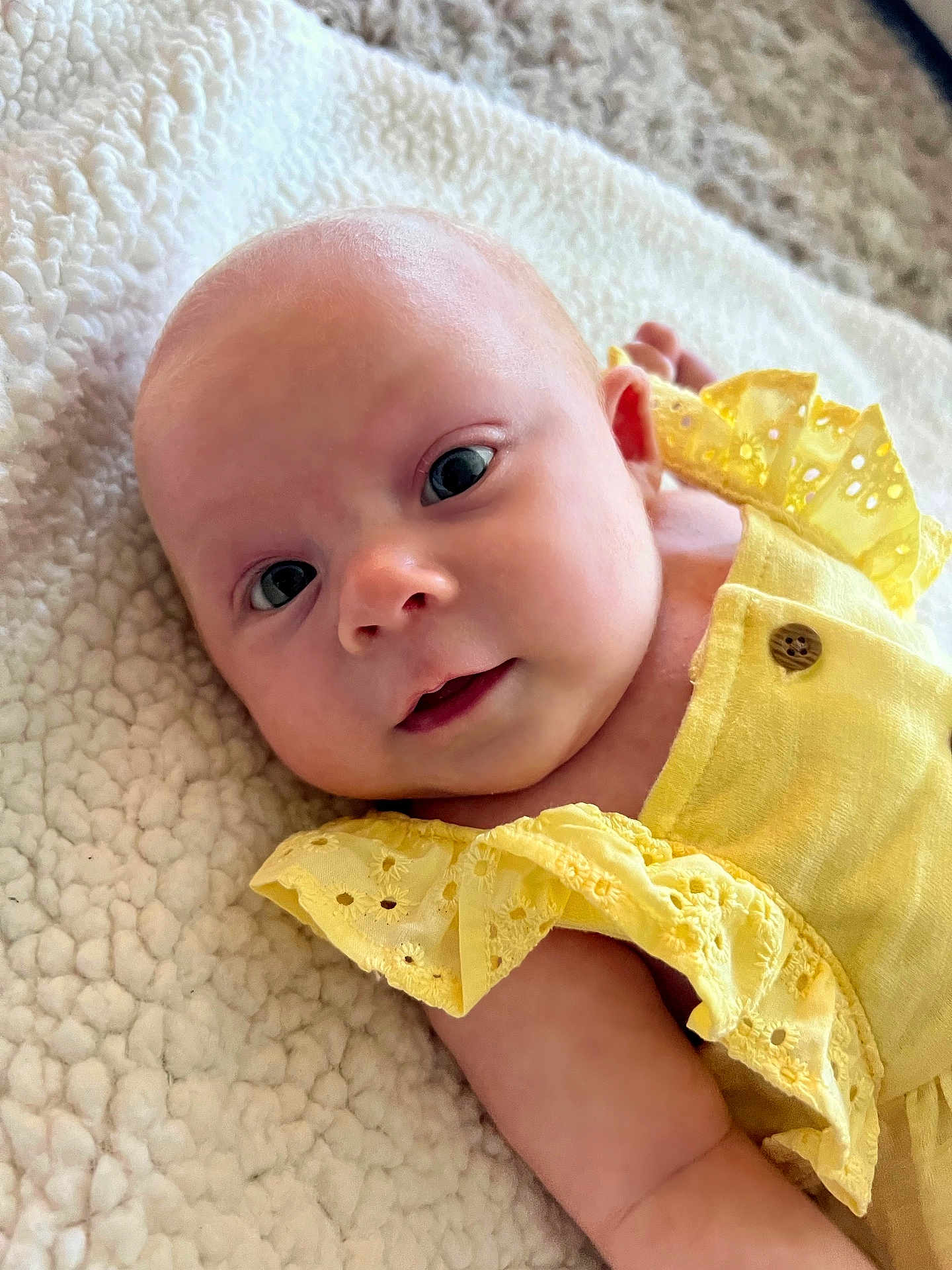 Bella is registered to the contest to win money with this photo: baby, infant, newborn, yellow_dress, ruffle, button, blanket, textured_blanket, closeup, face, eyes, gaze, cheek, skin, arm, portrait, soft_lighting, cozy, lying_down, adorable