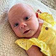 Bella is registered to the contest to win money with this photo: baby, infant, newborn, yellow_dress, ruffle, button, blanket, textured_blanket, closeup, face, eyes, gaze, cheek, skin, arm, portrait, soft_lighting, cozy, lying_down, adorable