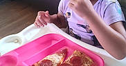 Jasabelle is registered to the contest to win money with this photo: toddler, child, curly_hair, high_chair, pizza, popcorn, chips, pink_tray, eating, snack, indoor, living_room, sofa, casual_clothing, food, meal_time, cute, young_child, portrait, happy