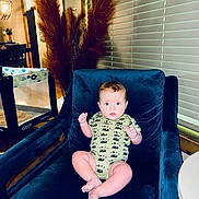 Silas is registered to the contest to win money with this photo: baby, child, blue_eyes, onesie, car_pattern, chair, plush, indoor, lamp, playpen, dried_plants, window_blinds, wood_ceiling, table, flooring, furniture, curious, seated, homedecor, person