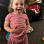 toddler, child, pink_clothing, shorts, tshirt, smiling, tongue_out, living_room, carpet, couch, dog, blanket, toy, earrings, person, happy, indoor, hair_tied, home, cute