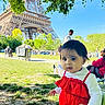 child, toddler, red_dress, outdoor, park, eiffel_tower, greenery, grass, sunny_day, people, adult, trees, sky, baby, nature, tourist_spot, relaxing, sitting, walking, shadows