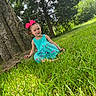 child, girl, grass, tree, nature, outdoor, greenery, smile, bow, dress, sunlight, park, young_child, happy, sitting, playful, casual, daylight, scenery, portrait