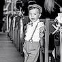 child, boy, vintage_clothing, suspenders, bow_tie, flat_cap, church, pew, aisle, flower_decorations, black_and_white, formal_event, leaning, casual_pose, smiling, shoes, floor, people, background, portrait