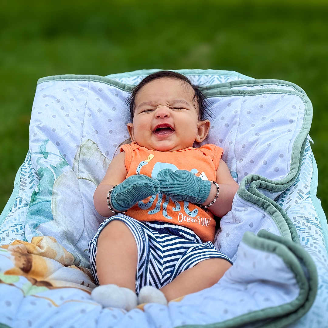 Reyansh is registered to the contest to win money with this photo: baby, blanket, child, cute, fabric, grass, green_background, happy, infant, laughing, mittens, orange_clothing, outdoor, patterned_blanket, person, seated, smiling, socks, striped_shorts, young_child