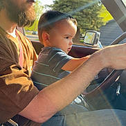 Bentley is registered to the contest to win money with this photo: child, adult, car, steering_wheel, sunlight, lens_flare, glasses, beard, cap, shirt, seatbelt, window, tree, side_mirror, portrait, inside_vehicle, casual_clothing, daylight, concentration, driving