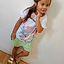 child, girl, standing, white_tshirt, green_skirt, sandals, wooden_floor, plain_wall, long_hair, necklace, casual_pose, smile, indoors, fashion, portrait, cute, young, person, footwear, hand_on_chin
