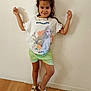 child, girl, smiling, standing, posing, indoor, white_wall, green_shorts, graphic_tshirt, sandals, wooden_floor, happy, casual_clothing, portrait, person, fashion, cute, playful, young, feet