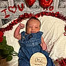 Amani is registered to the contest to win money with this photo: baby, infant, denim_overalls, smiling, raised_fist, red_decorations, heart_shapes, newborn, white_blanket, portrait, one_month, plant_leaves, background_photos, festive, cute, indoor, celebration, happy, red_garland, face