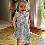 barefoot, blue_dress, casual, child, curly_hair, daisy_pattern, dress, hair_puffs, happy, home, indoor, natural_light, person, smile, standing, sunlight, tile_floor, toddler, window, young_child