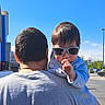 baby, child, toddler, sunglasses, outdoor, sunny, blue_sky, adult, parent, shoulder_ride, portrait, parking_lot, street, cars, building, traffic_sign, stop_sign, daylight, finger, casual_clothing