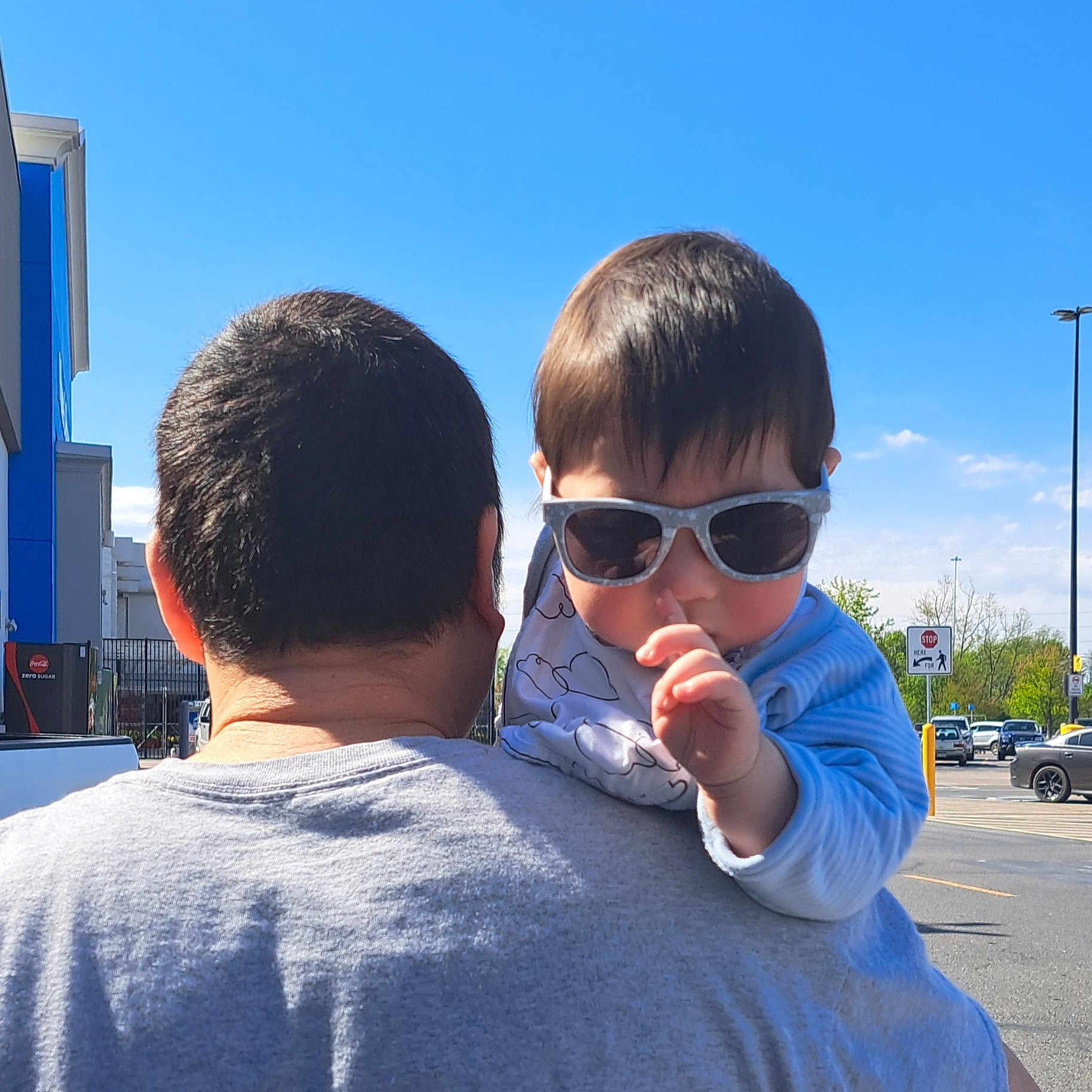 Viet is registered to the contest to win money with this photo: baby, child, toddler, sunglasses, outdoor, sunny, blue_sky, adult, parent, shoulder_ride, portrait, parking_lot, street, cars, building, traffic_sign, stop_sign, daylight, finger, casual_clothing