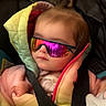 baby, child, puffer_jacket, car_seat, seatbelt, sunglasses, reflection, colorful_clothing, indoor, warm_clothing, cute, portrait, infant, young_child, face, head, hair_tie, pink, yellow, black