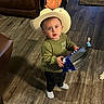 toddler, child, cowboy_hat, toy, indoor, wooden_floor, curious, standing, green_sweater, black_pants, socks, living_room, backpack, brown_couch, rug, flooring, playtime, cute, portrait, home