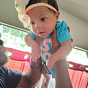 Tay'Lynn is registered to the contest to win money with this photo: baby, infant, headband, adult_hand, indoor, curtains, window, light, person, blue_clothing, pink_text, face, skin, arm, expression, holding, human, child, cute, happy