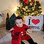 toddler, child, christmas_tree, holiday, gift_box, santa_costume, red_clothing, indoor, tiles, decorations, lights, shopping_bag, floor, festive, cute, present, celebration, playful, holiday_season, curious
