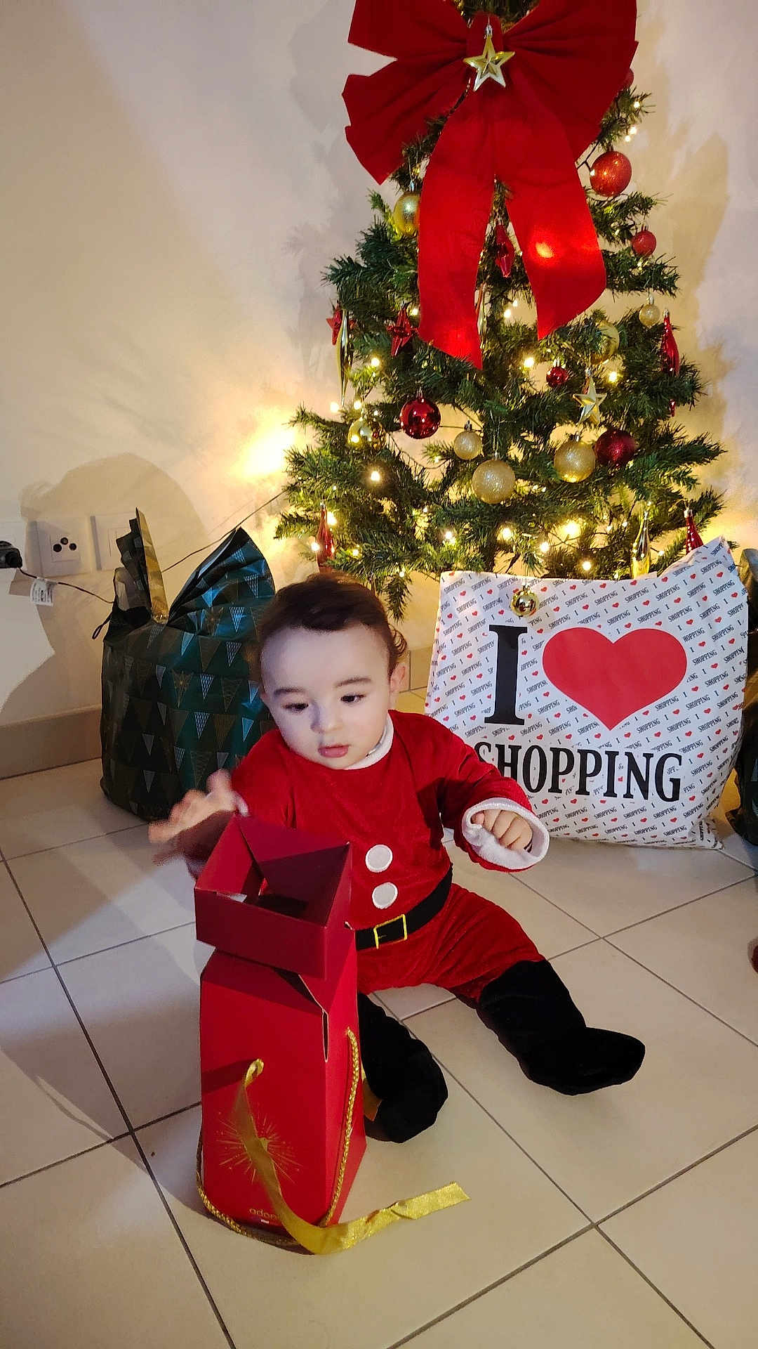 Ayden a rejoint le concours — aidez-le/la à gagner de superbes lots ! toddler, child, christmas_tree, holiday, gift_box, santa_costume, red_clothing, indoor, tiles, decorations, lights, shopping_bag, floor, festive, cute, present, celebration, playful, holiday_season, curious