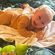 Austin joined the competition — help win amazing prizes! baby, smiling, onesie, blanket, bed, plush_toy, caterpillar_toy, green, brown, soft_texture, happy, infant, face, indoor, cute, child, lying_down, cozy, comfort, playful