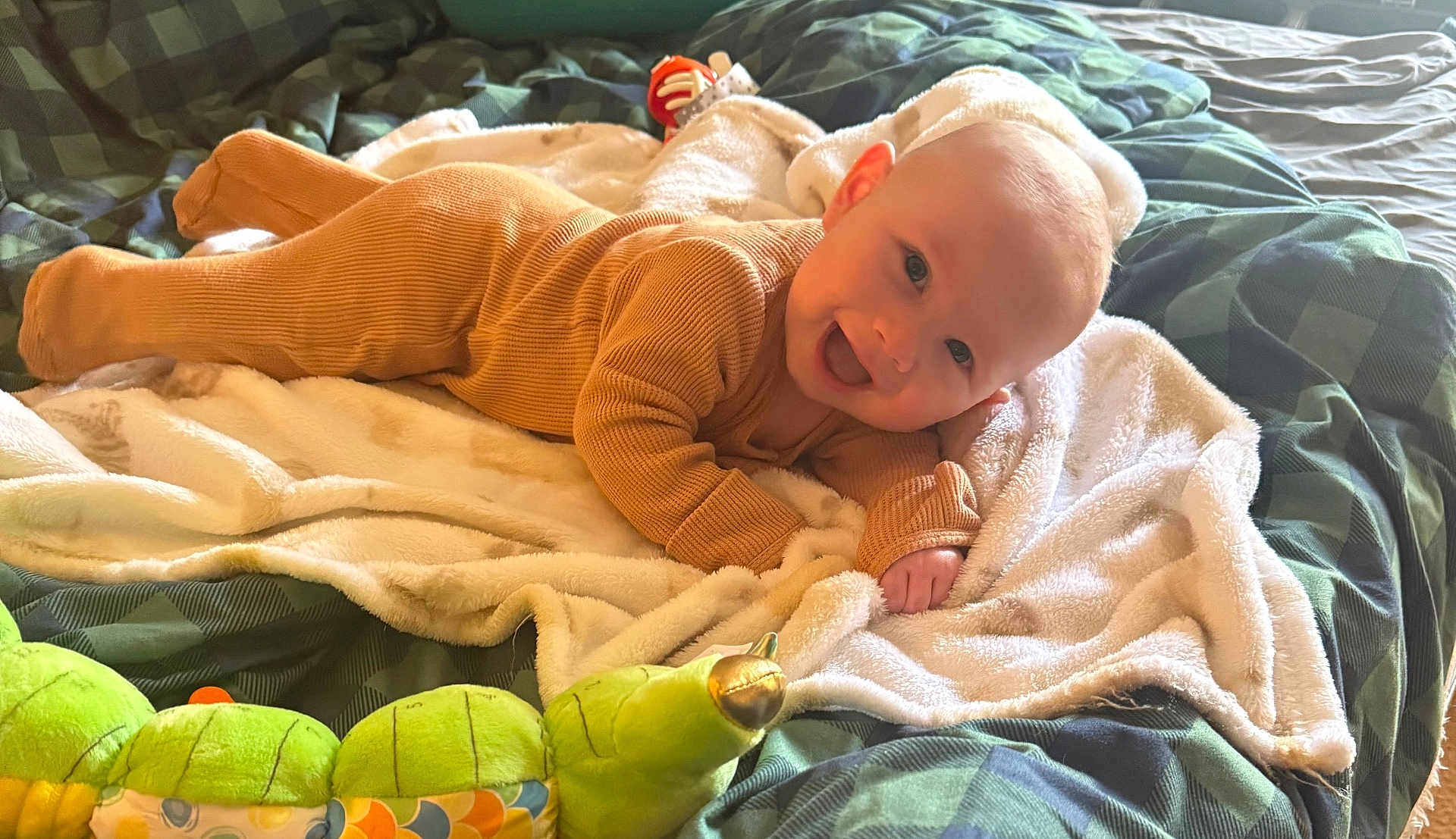 Austin joined the competition — help win amazing prizes! baby, smiling, onesie, blanket, bed, plush_toy, caterpillar_toy, green, brown, soft_texture, happy, infant, face, indoor, cute, child, lying_down, cozy, comfort, playful