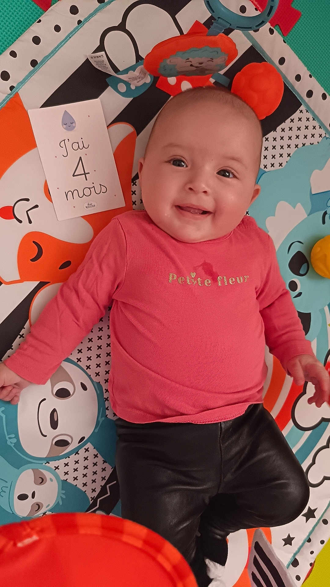 Hyuna a rejoint le concours — aidez-le/la à gagner de superbes lots ! baby, infant, smiling, playmat, milestone_card, french_text, pink_shirt, black_pants, face, eyes, tongue, toy, colorful, pattern, mat, portrait, laying, happy, closeup, baby_girl