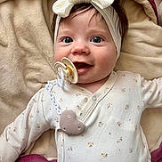 Mia is registered to the contest to win money with this photo: baby, infant, headband, bow, pacifier, onesie, blanket, smile, face, child, cute, happy, lying_down, apparel, clothing, soft_texture, indoors, portrait, closeup, baby_girl