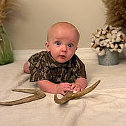 Henry is registered to the contest to win money with this photo: baby, child, infant, camouflage_clothing, antlers, curious_expression, lying_down, blanket, vase, cotton_stems, homedecor, indoor, person, portrait, soft_lighting, face, hands, floor, cute, toddler
