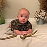 baby, child, infant, camouflage_clothing, antlers, curious_expression, lying_down, blanket, vase, cotton_stems, homedecor, indoor, person, portrait, soft_lighting, face, hands, floor, cute, toddler
