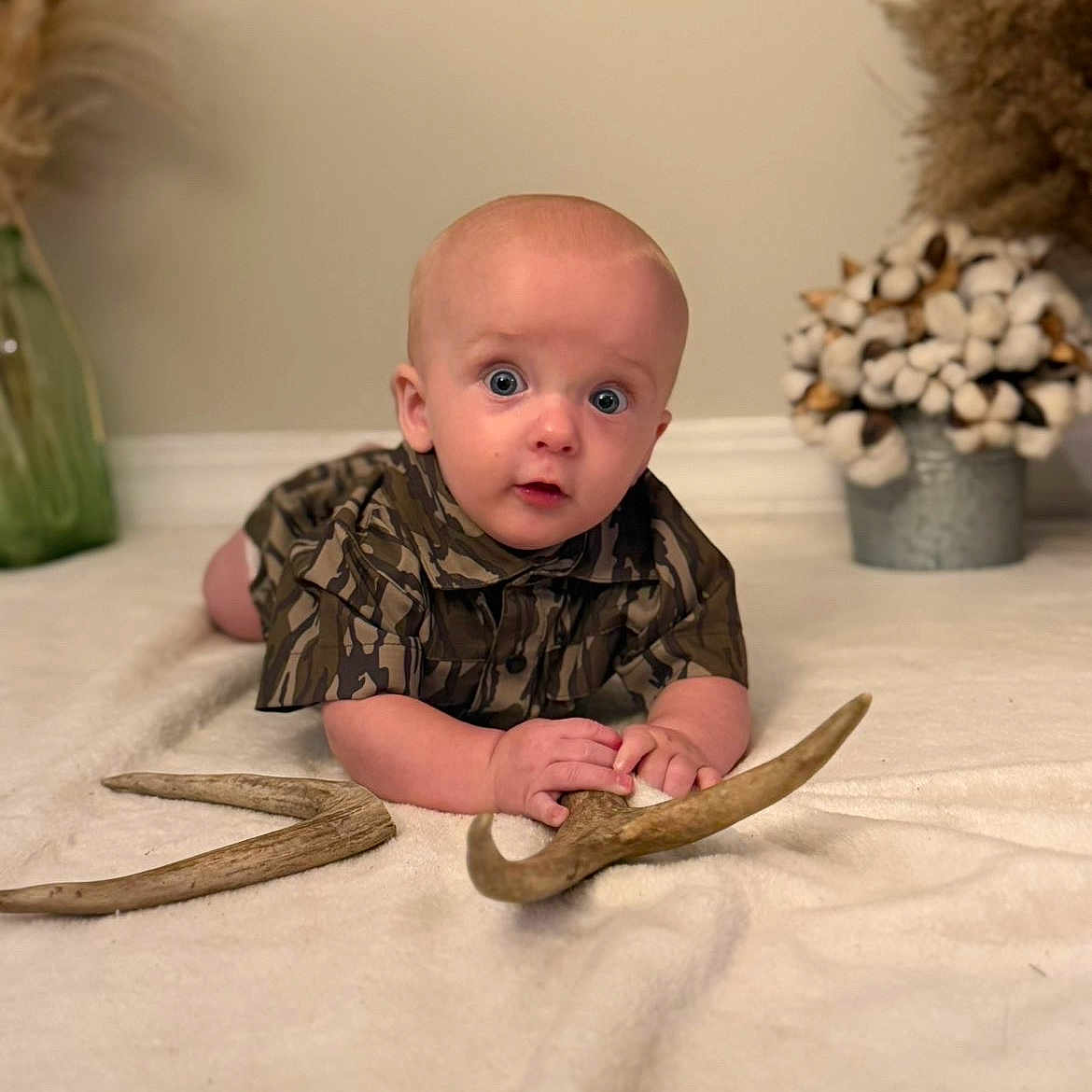 Henry is registered to the contest to win money with this photo: baby, child, infant, camouflage_clothing, antlers, curious_expression, lying_down, blanket, vase, cotton_stems, homedecor, indoor, person, portrait, soft_lighting, face, hands, floor, cute, toddler