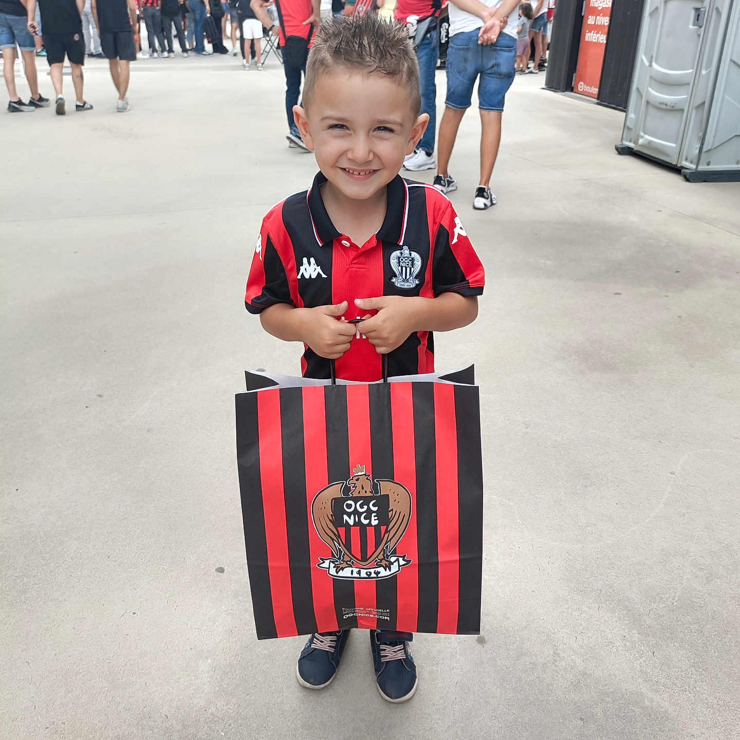 Myliano a rejoint le concours — aidez-le/la à gagner de superbes lots ! boy, casual_clothing, child, concrete, crowd, daytime, event, hair_spiked, happy, ogc_nice, outdoor, people, portrait, red_and_black, shoes, shopping_bag, shorts, smiling, sports_jersey, standing