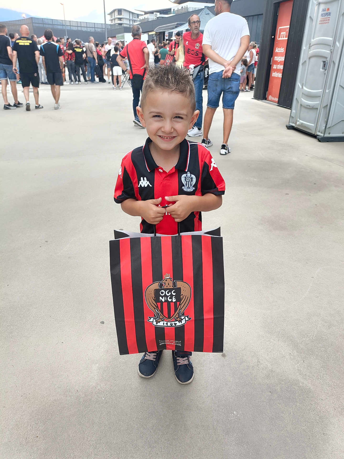 Myliano a rejoint le concours — aidez-le/la à gagner de superbes lots ! child, boy, smiling, shopping_bag, red_and_black, sports_jersey, ogc_nice, crowd, outdoor, concrete, people, casual_clothing, shorts, shoes, daytime, event, hair_spiked, standing, portrait, happy