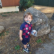 Andrés a rejoint le concours — aidez-le/la à gagner de superbes lots ! blue_clothing, building, child, daylight, ground, happy, leafy, maroon_clothing, number_10, outdoor, pacifier, person, plants, rock, shirt, shorts, smiling, sneakers, soccer_outfit, toddler